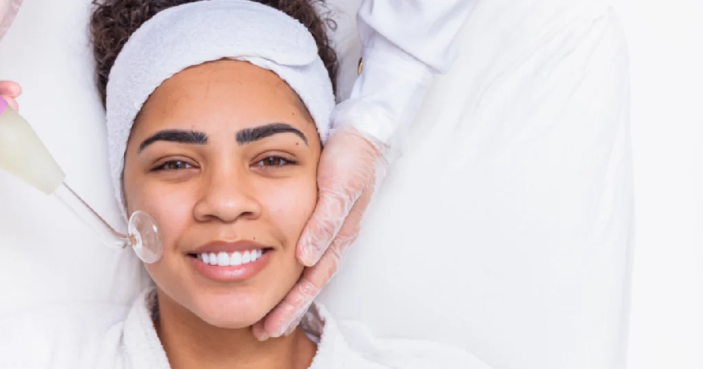 A smiling woman lies on a white cushion while a person in white gloves performs a Medical Grade Skincare facial on her using a cosmetic tool in Scottsdale, AZ