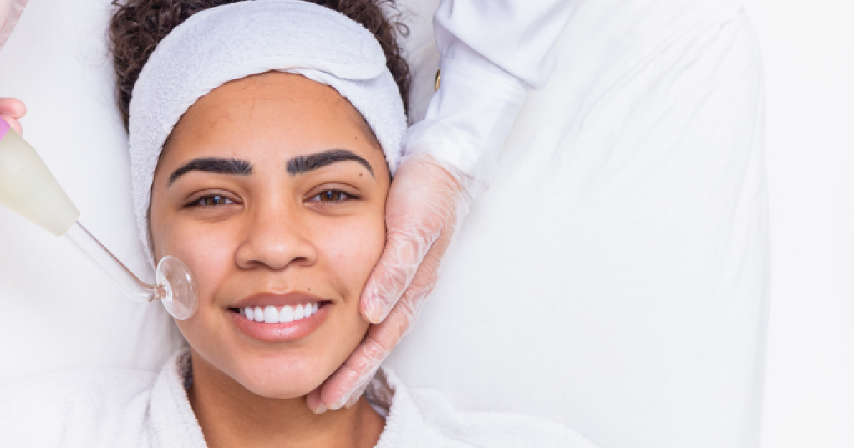A smiling woman lies on a white cushion while a person in white gloves performs a Medical Grade Skincare facial on her using a cosmetic tool in Scottsdale, AZ